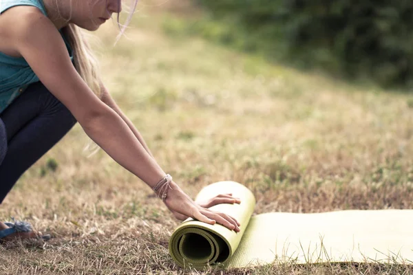 Cropped image of girl rolling yoga mat. Close-up of attractive young woman folding green yoga or fitness mat after working out in a glade in the woods or a city Park . Healthy life