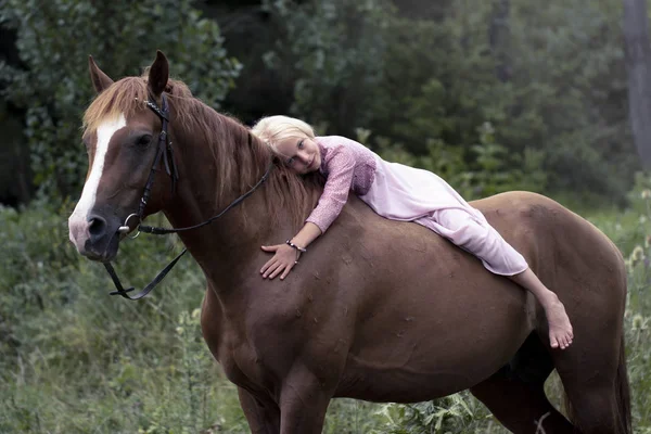 blonde girl with horse in the forest. beautiful Caucasian girl with long blonde hair in a pink dress lies on a brown horse and hugs her. barefoot child on horseback in the forest Bareback.