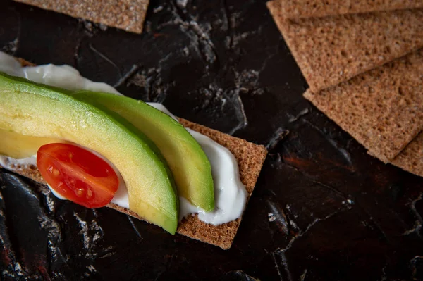 Snack saludable de galleta de pan crujiente de centeno integral con tomates Cherry, aguacate y yogur sobre fondo negro — Foto de Stock