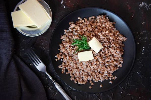 Porridge di grano saraceno appena cotto con pezzi di burro in un piatto nero su sfondo scuro. Vista dall'alto. Cibo sano. — Foto Stock