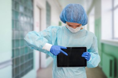 Koruyucu giysili kadın doktor: tıbbi maske, şapka ve bornoz. Kadın dijital tablet kullanıyor ve ekranda hastalık istatistikleri gösteriyor..