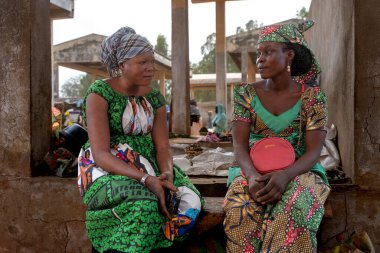 Geleneksel elbiseli iki kadın, günlük yaşamı ve toplumun coşkulu ruhunu yansıtan bir Kamerun pazarında sohbet ediyorlar..