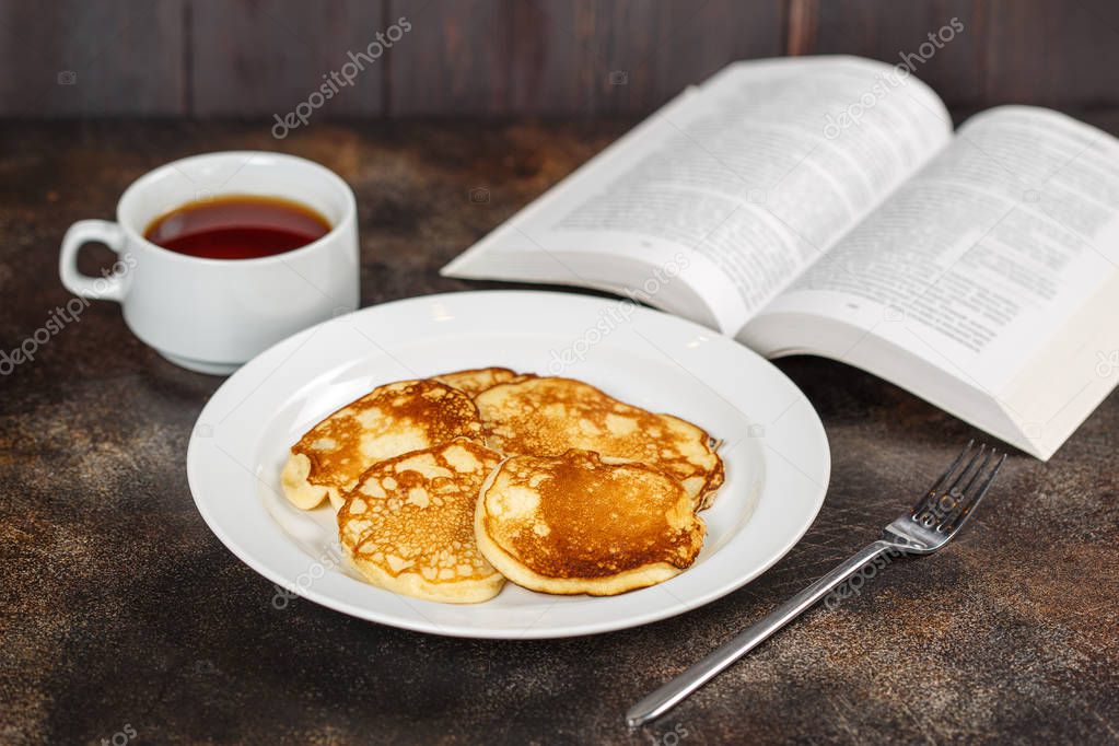 Pancakes on white plate with tea and book on brown background