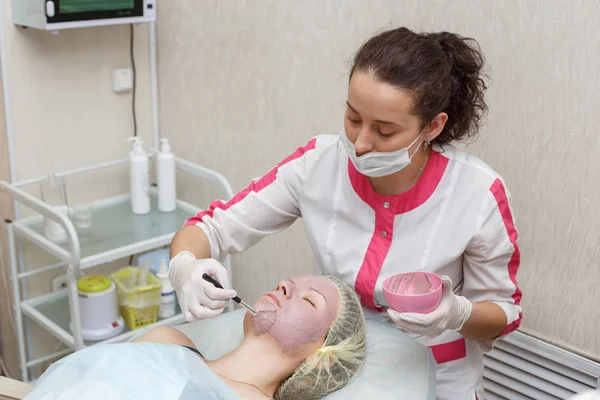 Woman getting pink face peeling mask in spa beauty salon by beautician
