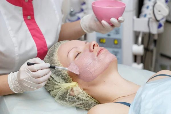 Woman getting pink face peeling mask in spa beauty salon by beautician