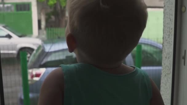 Un niño se para cerca de una ventana y mira la lluvia en la calle. La lluvia deja de jugar — Vídeos de Stock