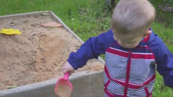 Ein kleiner Junge spielt mit einem Sandkasten und wischt Sand von den Turnschuhen. — Stockvideo