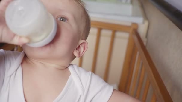 Двухлетний мальчик пьет молоко из пластиковой бутылки в своей постели, глядя вверх — стоковое видео