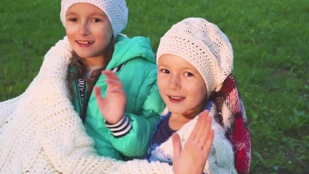 Две счастливые улыбающиеся девушки сидят в парке и машут руками перед камерой. Дети на фоне цветущего сада . — стоковое видео