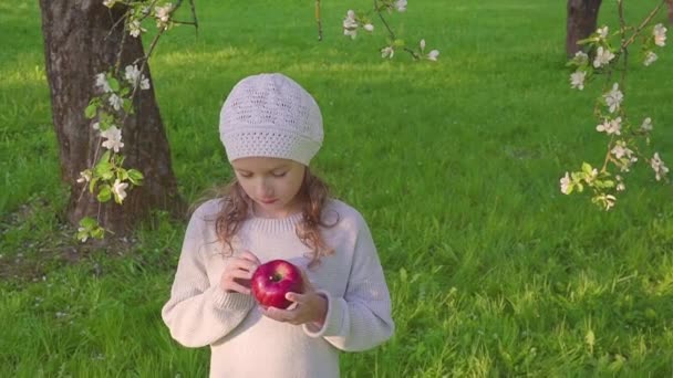 Kleine meisje portret rode appel buiten eten. Een meisje met een appel op de achtergrond van de bloeiende tuinen. — Stockvideo