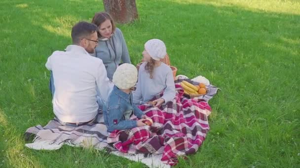 Família alegre sentada na grama durante um piquenique em um parque, todos tomam café da manhã. há um cesto com refeição. Frutos frescos — Vídeo de Stock