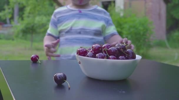 Menino a comer cerejas no jardim. Cerejas em uma chapa em uma mesa . — Vídeo de Stock