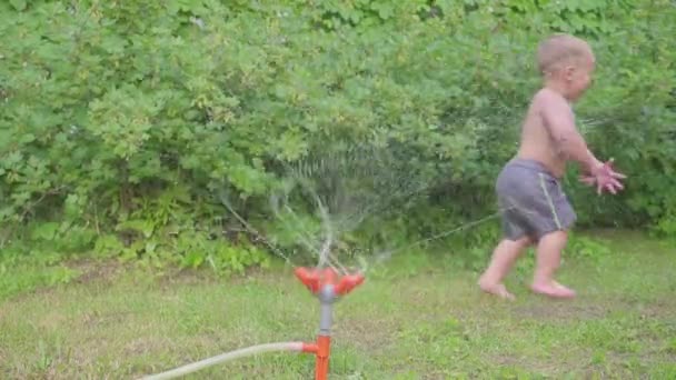 Счастливый мальчик веселится на улице с водяным разбрызгивателем в летнем саду. Медленное движение — стоковое видео
