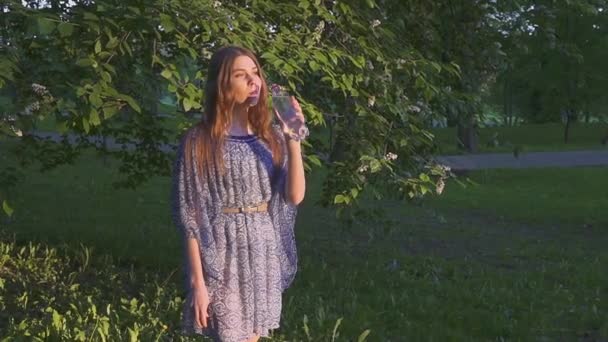 Beautiful female hiker drinking water in forest at sunset. A girl in a dress drinks cool water from a plastic bottle. — Stock Video