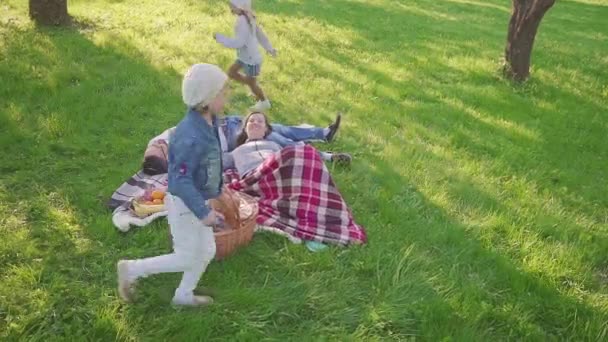 Szülők feküdt a fűben, hogy két lánya van szórakoztató és futás háttérben park. Családi nyaralás a táj. — Stock videók