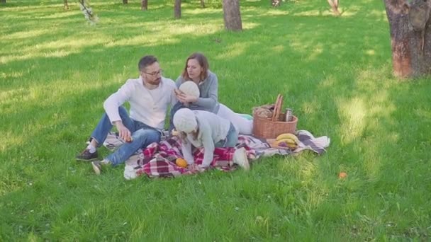 Família alegre sentada na grama durante um piquenique em um parque, todos tomam café da manhã. há um cesto com refeição. Frutos frescos — Vídeo de Stock