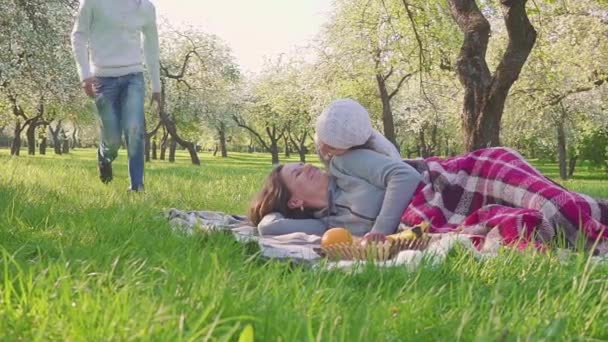 La familia feliz pasa tiempo en un picnic cerca del manzano floreciente. La madre yace en la hierba, los niños y el marido corren hacia ella. Todo el mundo besa madre . — Vídeo de stock