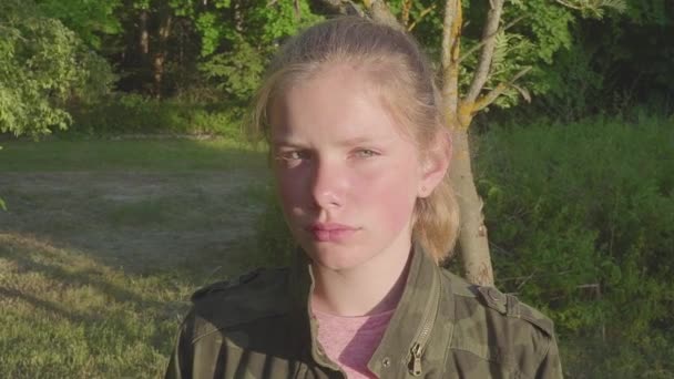 Portrait rapproché d'une jolie adolescente triste regardant la caméra. Enfant souriant heureux blanc aux cheveux longs blonds naturels debout à l'extérieur . — Video