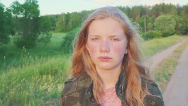 Ein verträumtes Teenager-Mädchen in Militärjacke, das auf einem Feld in der Nähe eines Waldes in der Natur spaziert. das Kind hat blonde Haare. — Stockvideo