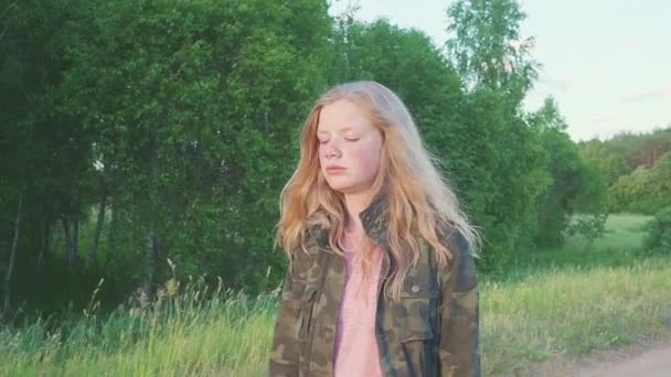 自然の中の森付近フィールドを歩いてミリタリー ジャケットで夢のような悲しい十代の少女。その子はブロンドの髪。ステディカム撮影. — ストック動画