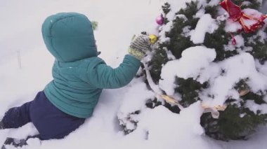 çocuk çocuk Noel ağacı yakınındaki kar üzerinde yatıyor ve açık süslemeleri ile oynanır.