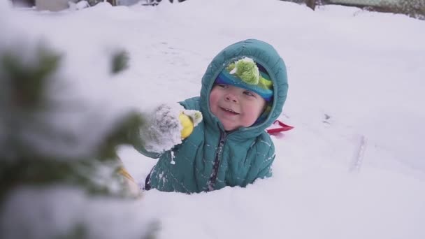 Мальчик ребенок лежит на снегу рядом с елкой и играет с украшениями на открытом воздухе . — стоковое видео