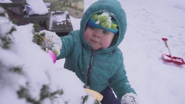 Ragazzo il bambino si trova sulla neve vicino all'albero di Natale ed è giocato con decorazioni all'aperto . — Video Stock