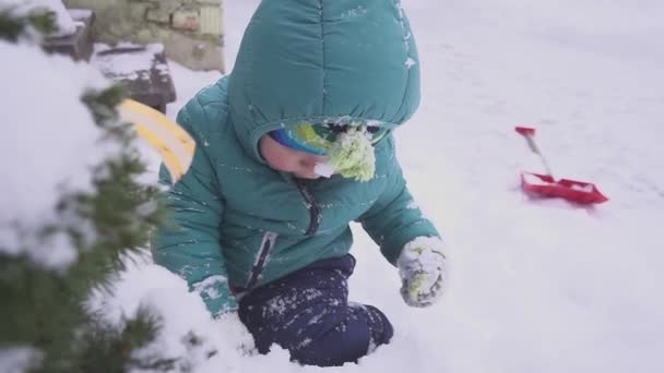 Αγόρι το παιδί βρίσκεται στο χιόνι κοντά το χριστουγεννιάτικο δέντρο και παίζεται με υπαίθρια διακοσμήσεις. — Αρχείο Βίντεο