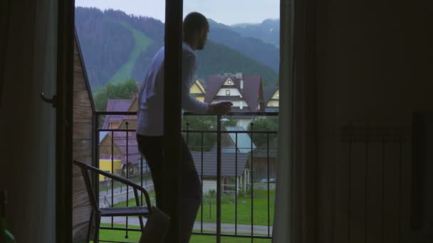Młody człowiek idzie z jego pokoju na balkon przy lampce wina, poszukaj w górach. — Wideo stockowe