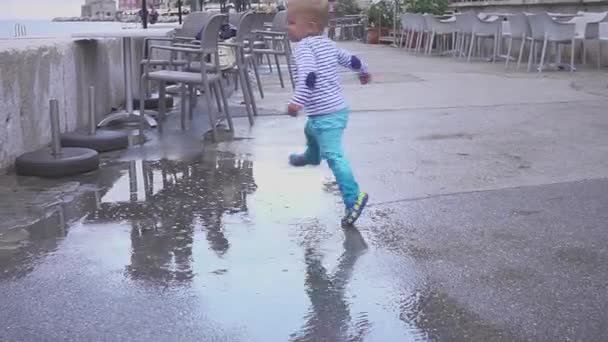 El chico está jugando en un charco bajo la lluvia. Se regocija en la lluvia. Niño de dos años corriendo en la acera . — Vídeos de Stock