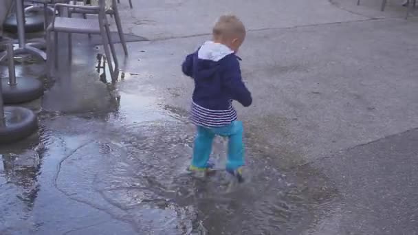 El chico está jugando en un charco bajo la lluvia. Se regocija en la lluvia. Niño de dos años corriendo en la acera . — Vídeos de Stock