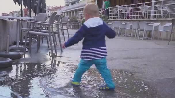 Le garçon joue dans une flaque sous la pluie. Il se réjouit de la pluie. Garçon de deux ans courant sur le trottoir . — Video