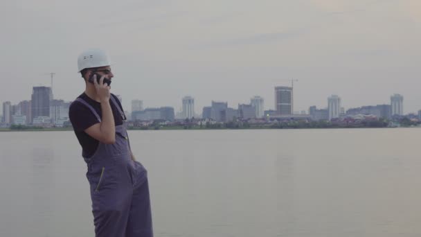 4K знімок красивого кавказького працівника в білому шоломі розмовляє на мобільному телефоні біля красивого вечірнього міста і будівельного майданчика. успіх будівництва — стокове відео
