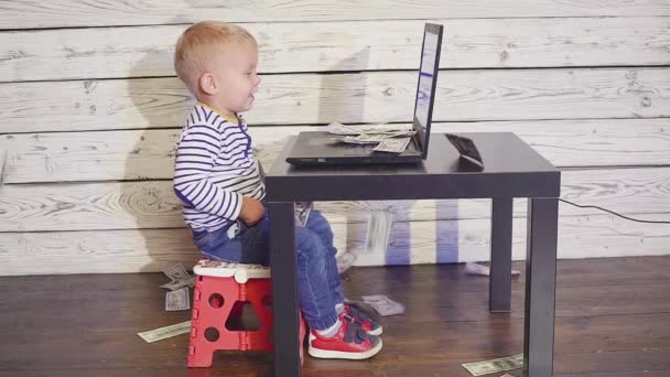 Дворічний хлопчик з комп'ютером і падаючими грошима. усміхнений хлопчик сидить за столом з ноутбуком і доларовими банкнотами в повітрі. Концепція успішного бізнесу — стокове відео