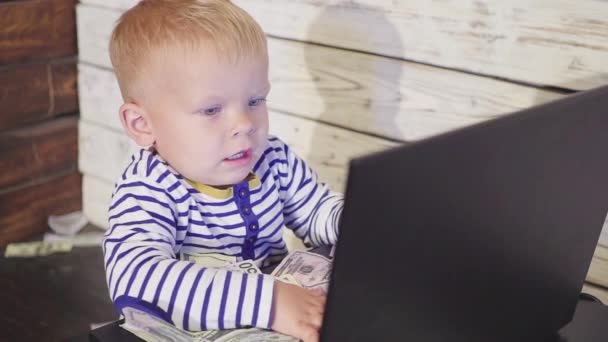 两岁的男孩拿着电脑, 钱也掉了。坐在桌子上的微笑的男孩与笔记本电脑和美元钞票在空气中。成功经营理念 — 图库视频影像