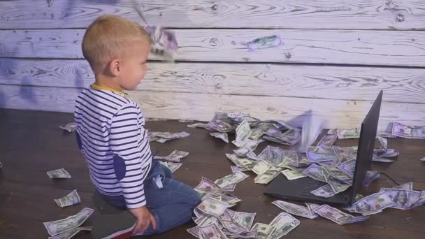 Két éves fiú egy számítógép és a csökkenő pénz. a laptop és a dollár bankjegyek levegőben asztalnál ülő fiú mosolyogva. A sikeres üzleti koncepció — Stock videók