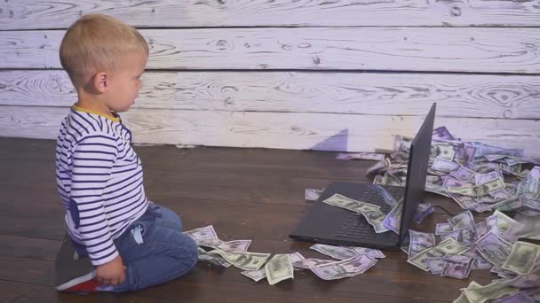 Bir bilgisayar ve düşen para ile iki yaşında bir çocuk. dizüstü bilgisayar ve doları banknot hava ile masada oturan boy gülümseyerek. Başarılı iş kavramı — Stok video