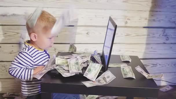 Két éves fiú egy számítógép és a csökkenő pénz. a laptop és a dollár bankjegyek levegőben asztalnál ülő fiú mosolyogva. Sikeres üzleti koncepciót. lassú mozgás — Stock videók