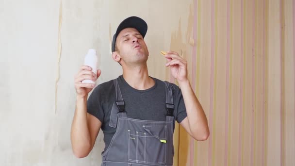 Builder eller byggande arbetare med nöje drycker från en vit flaska och äta en kaka inne i byggnaden i reparationer — Stockvideo