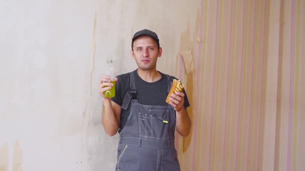 Builder or construction worker with pleasure drinks from a bottle and eating a cake inside in repairs building — Stock Video