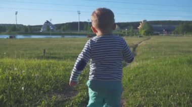 Mutlu bir çocukluk çalışan kamera için yeşil çim ve gülümseyerek. Küçük çocuk bir şehir parkında bir taze çimenlerin üzerinde ilk adımlarını alır. Beyaz dandelions alanında