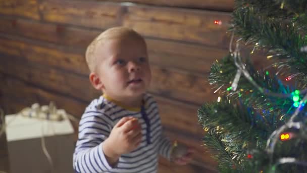 Zweijähriger Junge spielt mit Weihnachtsspielzeug am Weihnachtsbaum, aus nächster Nähe. Porträt eines Kindes neben einem Weihnachtsbaum. — Stockvideo