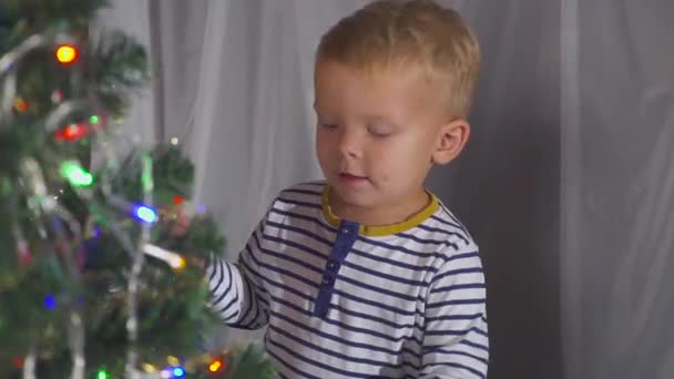 Zweijähriger Junge spielt mit Weihnachtsspielzeug am Weihnachtsbaum, aus nächster Nähe. Porträt eines Kindes neben einem Weihnachtsbaum. — Stockvideo