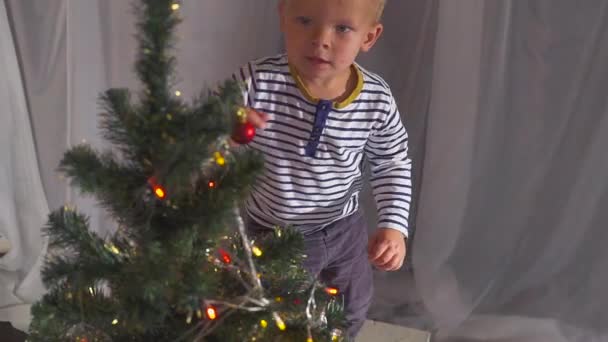 Two year old boy playing with Christmas toys on Christmas tree, close up. Portrait of a child near a Christmas tree. — Stock Video
