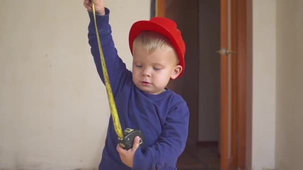 Lilla builder i röd hjälm med gula linjal tejp i händer i lägenheten under reparation. Konstruktion koncept. — Stockvideo