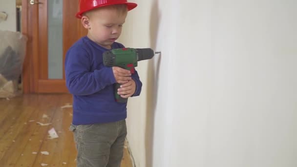 Young cute builder boy in uniform and hard hat or helmet with electric screwdriver against the background of construction and repair. — Stock Video