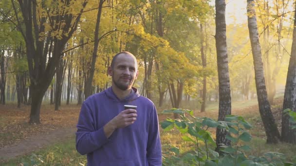 Молодий чоловік п'є чай або каву з паперової чашки в осінньому парку — стокове відео
