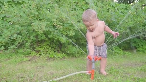 Милый мальчик веселится на улице с водяным разбрызгивателем в летнем саду. Медленное движение — стоковое видео