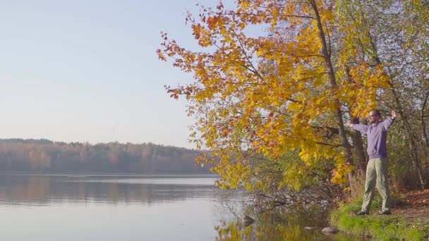 一个穿着蓝色毛衣的男人挥舞着他的手, 在秋天森林的湖面上展示了成功。. — 图库视频影像