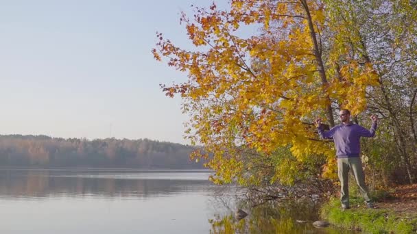 Egy férfi egy kék pulóver, intett a kezével, sikerét bizonyítja az őszi erdő a tavon. — Stock videók